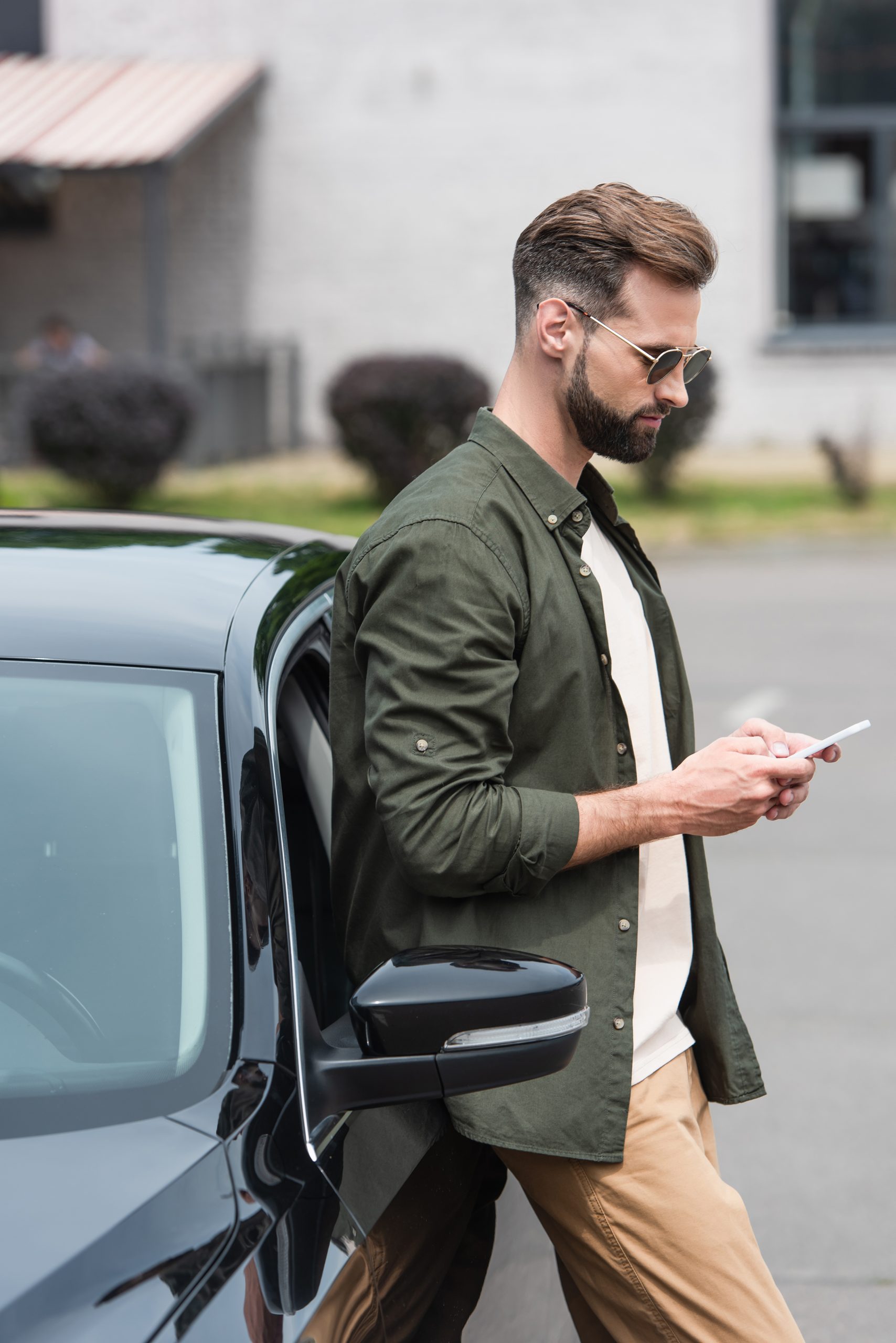 Side view of man in sunglasses using smartphone near car